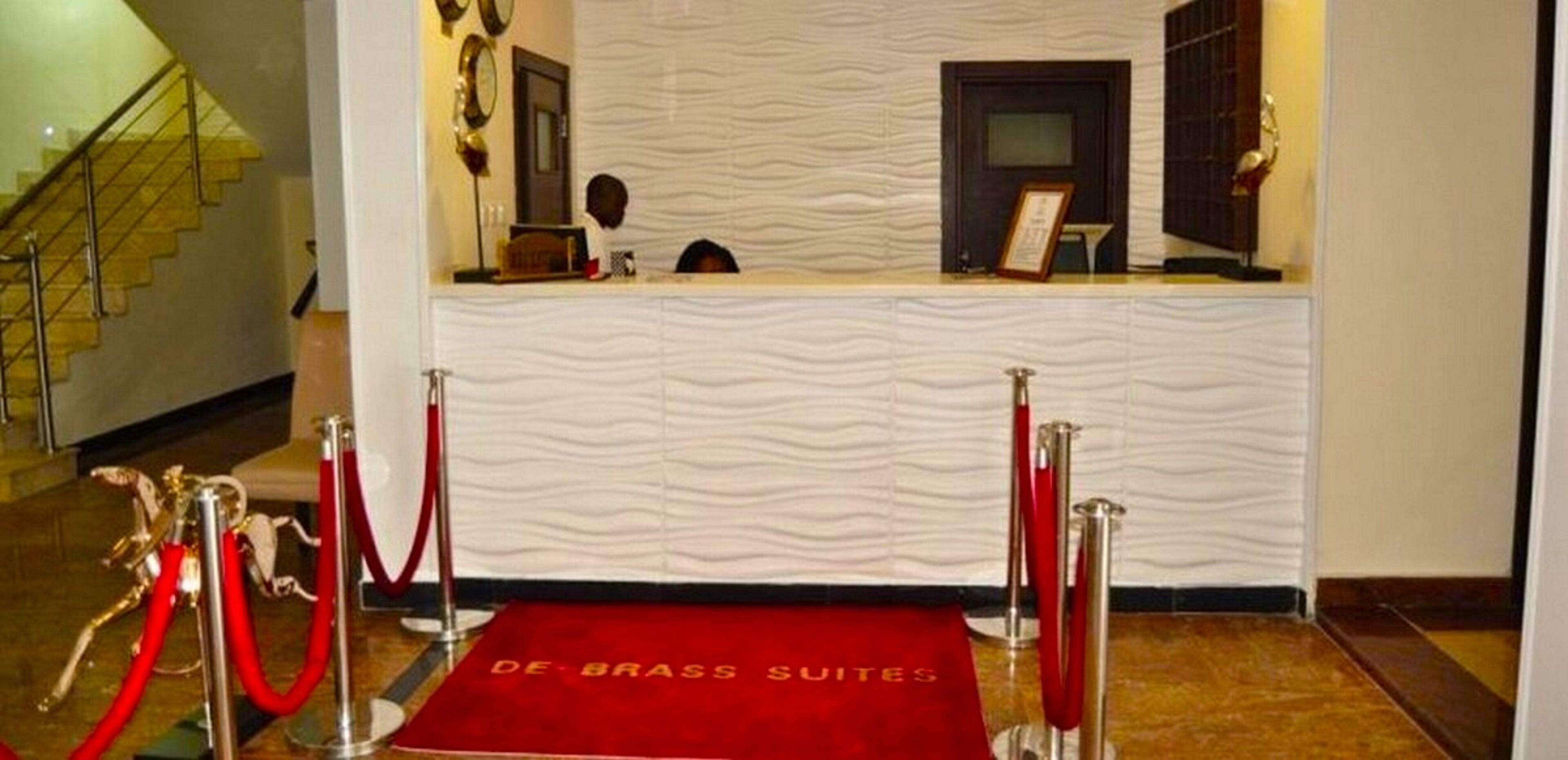 A reception area with a wavy textured wall, a red carpet with 'DE BRASS SUITES' text, and a staircase in the background.