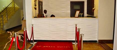 A reception area with a wavy textured wall, a red carpet with 'DE BRASS SUITES' text, and a staircase in the background.