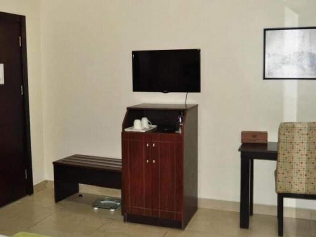A room with a television mounted on the wall, a small cabinet with a tray, a bench, and a chair.