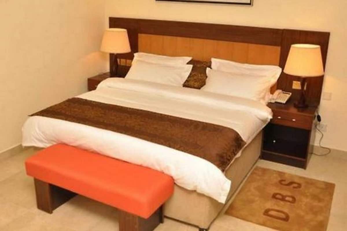 A neatly made bed with a brown blanket, two bedside lamps, a small bedside table, and a bench at the foot of the bed.