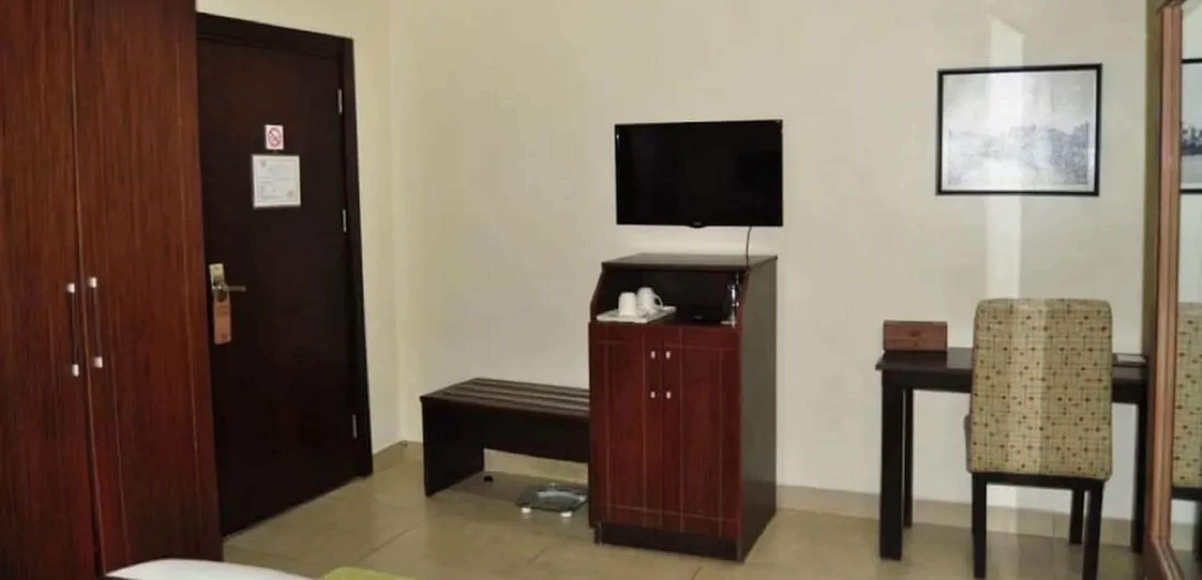 A room with a wooden door, a bench, a television on a stand, a chair, and a framed picture on the wall.