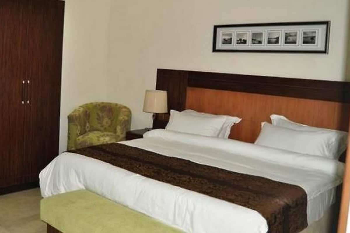 A hotel room with a large bed, a wooden headboard, a green armchair, a bedside table with a lamp, and a framed picture on the wall.