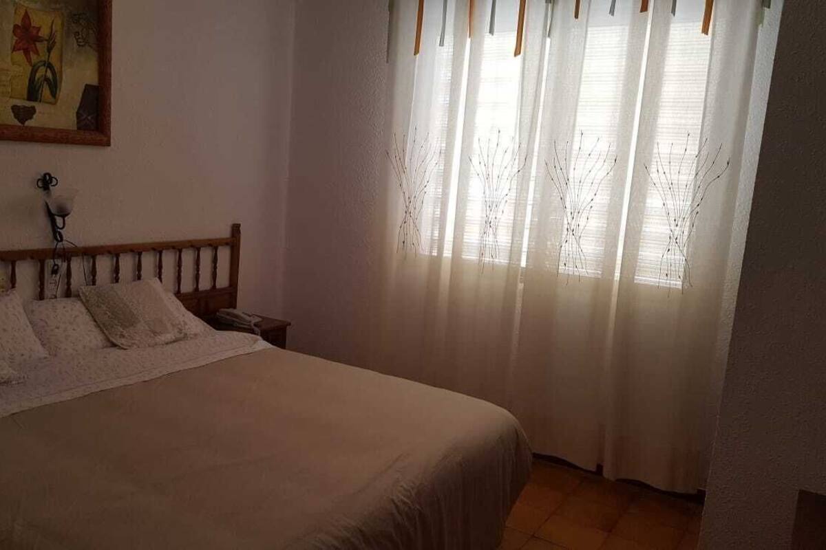 Een slaapkamer met een bed, een nachtkastje, een wandlamp en een raam met lichte gordijnen.