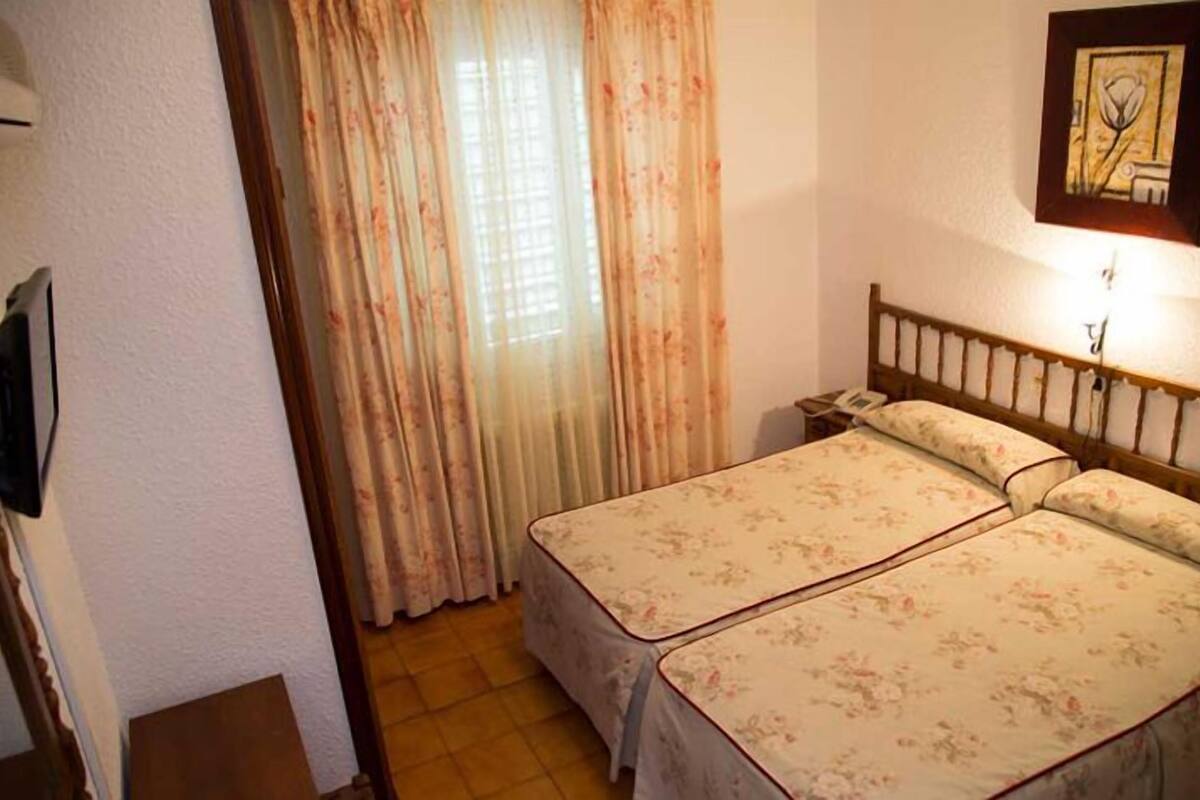 Een kamer met twee bedden, een houten hoofdbord, een nachtkastje, een wandtelefoon en een raam met gordijnen.