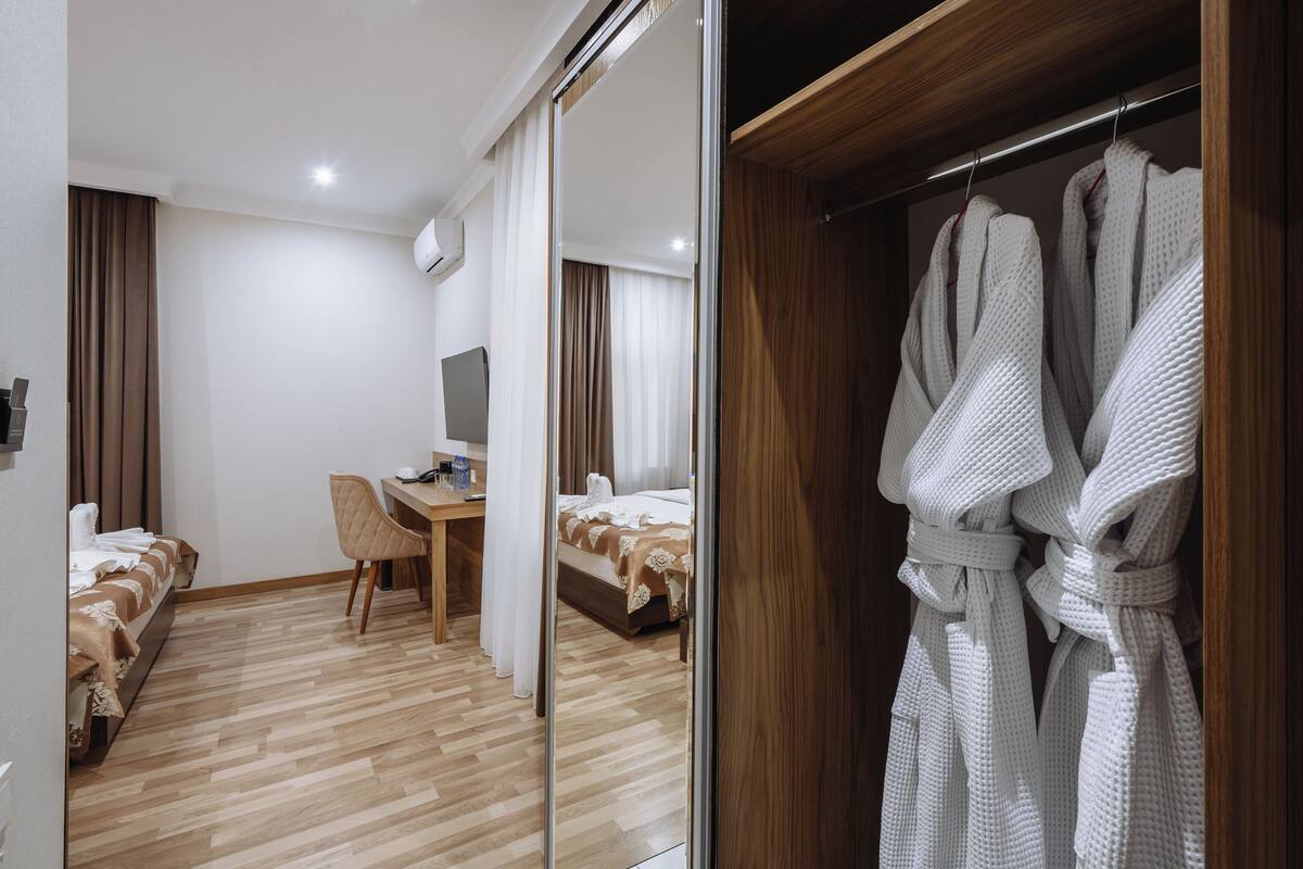 Une chambre d’hôtel avec un plancher en bois, un bureau avec une chaise, un lit recouvert d’une couverture blanche et un placard contenant des peignoirs blancs.