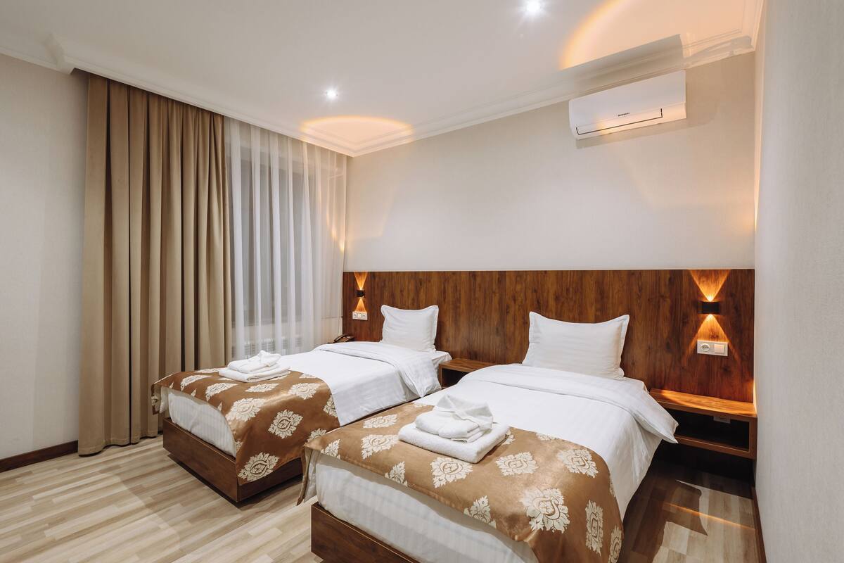 Une chambre d’hôtel avec deux lits, une tête de lit en bois, du linge de lit blanc et une grande fenêtre avec des rideaux.