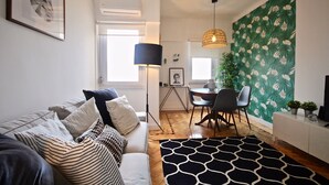 Una sala de estar moderna con un sofá, una mesa de comedor y sillas. Se observa un tapete con diseño blanco y negro, una pared con papel tapiz floral y una lámpara colgante.