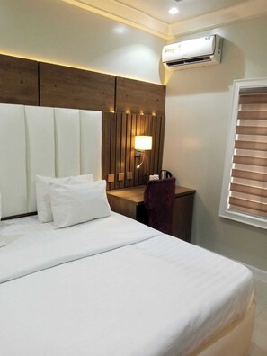 A hotel room with a bed, a nightstand, a lamp, an air conditioner, and a window with blinds.