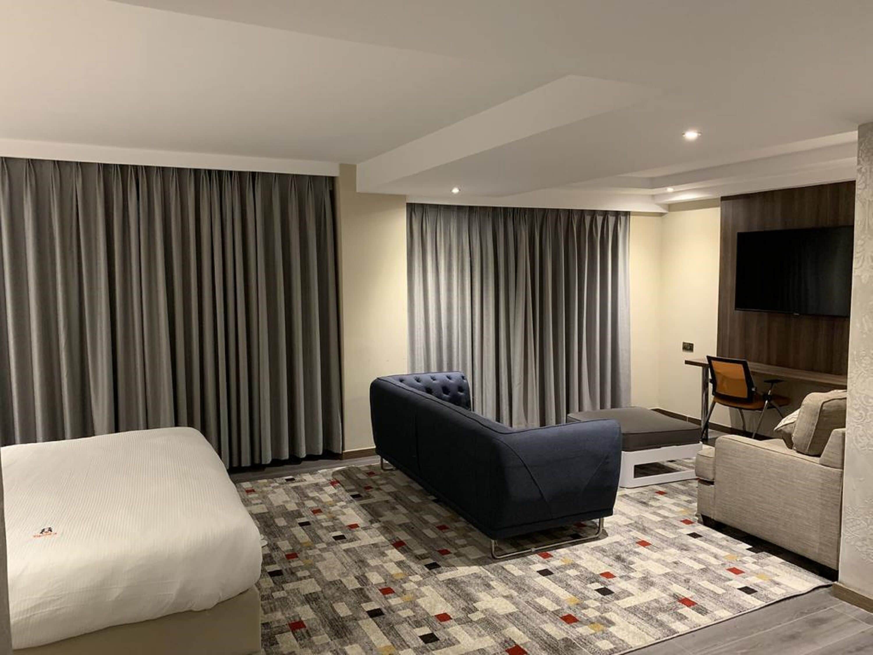 A hotel room with a large bed, a sofa, a blue armchair, a TV mounted on the wall, and a patterned rug.