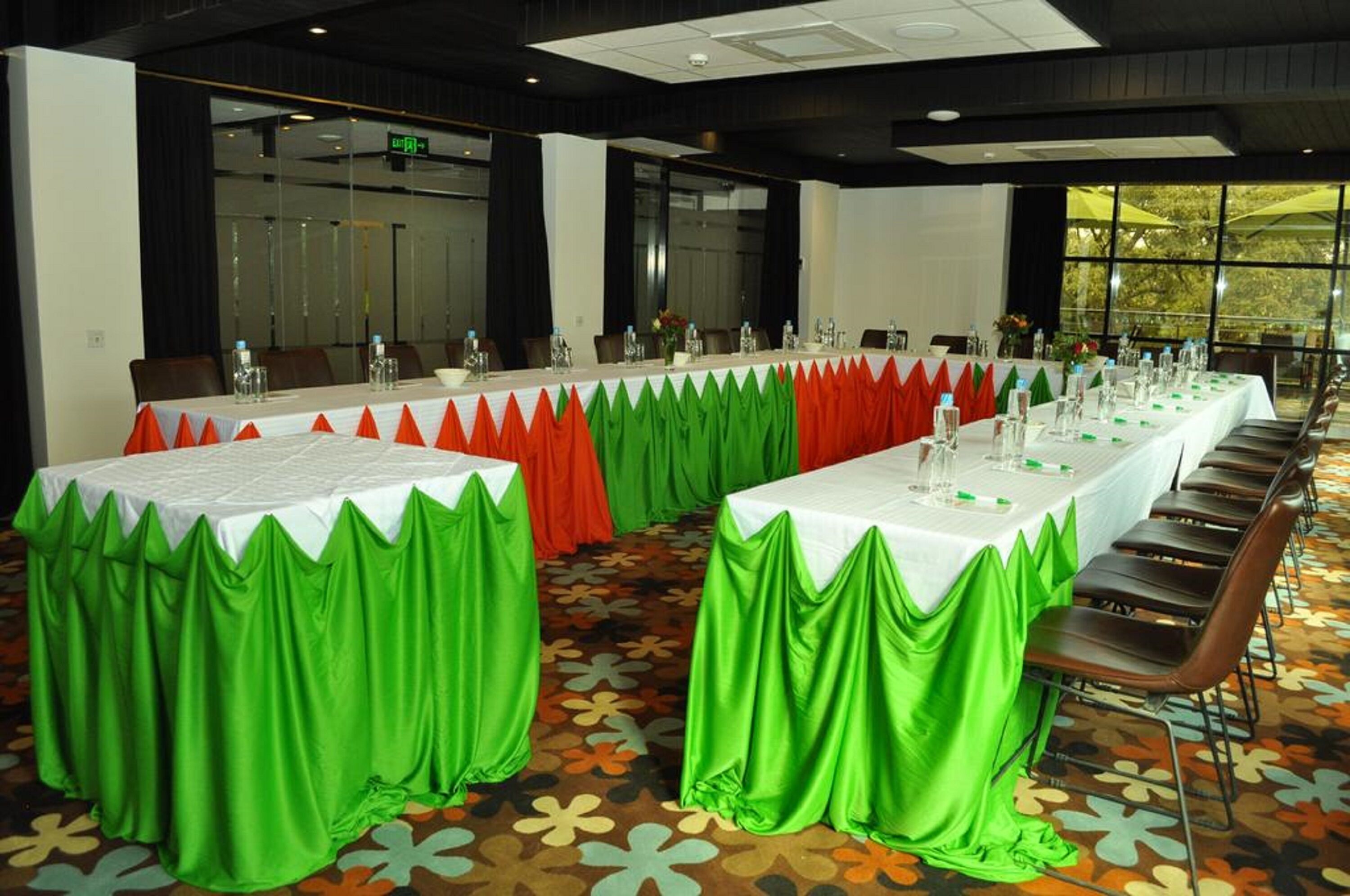 Een vergaderzaal met lange tafels, groene en rode gordijnen en een kamerbreed tapijt met een patroon.