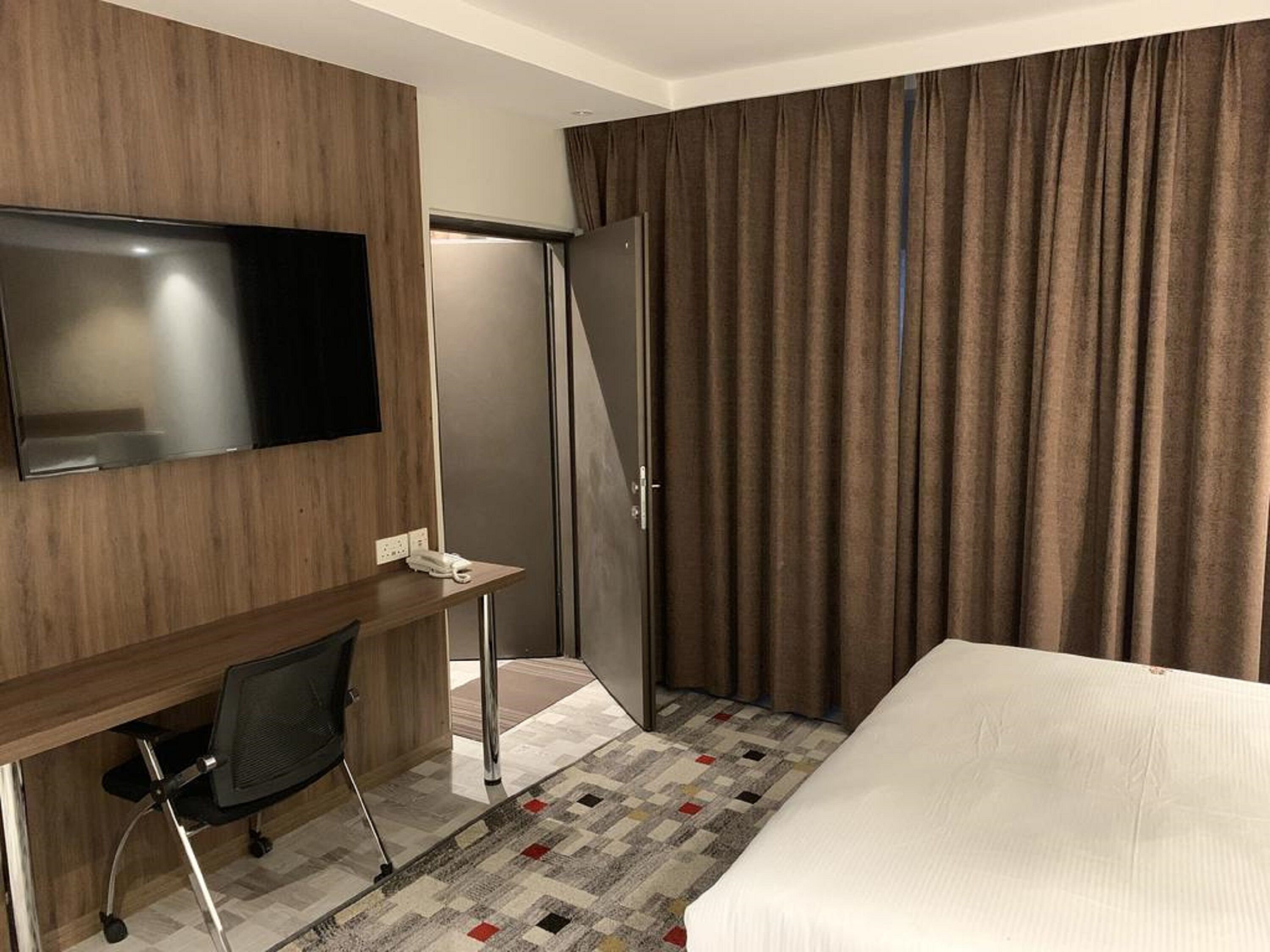 A hotel room with a bed, a desk with a chair, a flat-screen TV, a wardrobe, and a large window with curtains.