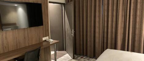 A hotel room with a bed, a desk with a chair, a flat-screen TV, a wardrobe, and a large window with curtains.