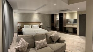 Una habitación de hotel moderna con una cama grande, un sofá con cojines estampados, una mesita de noche con lámparas, un armario y un espejo sobre el lavabo.