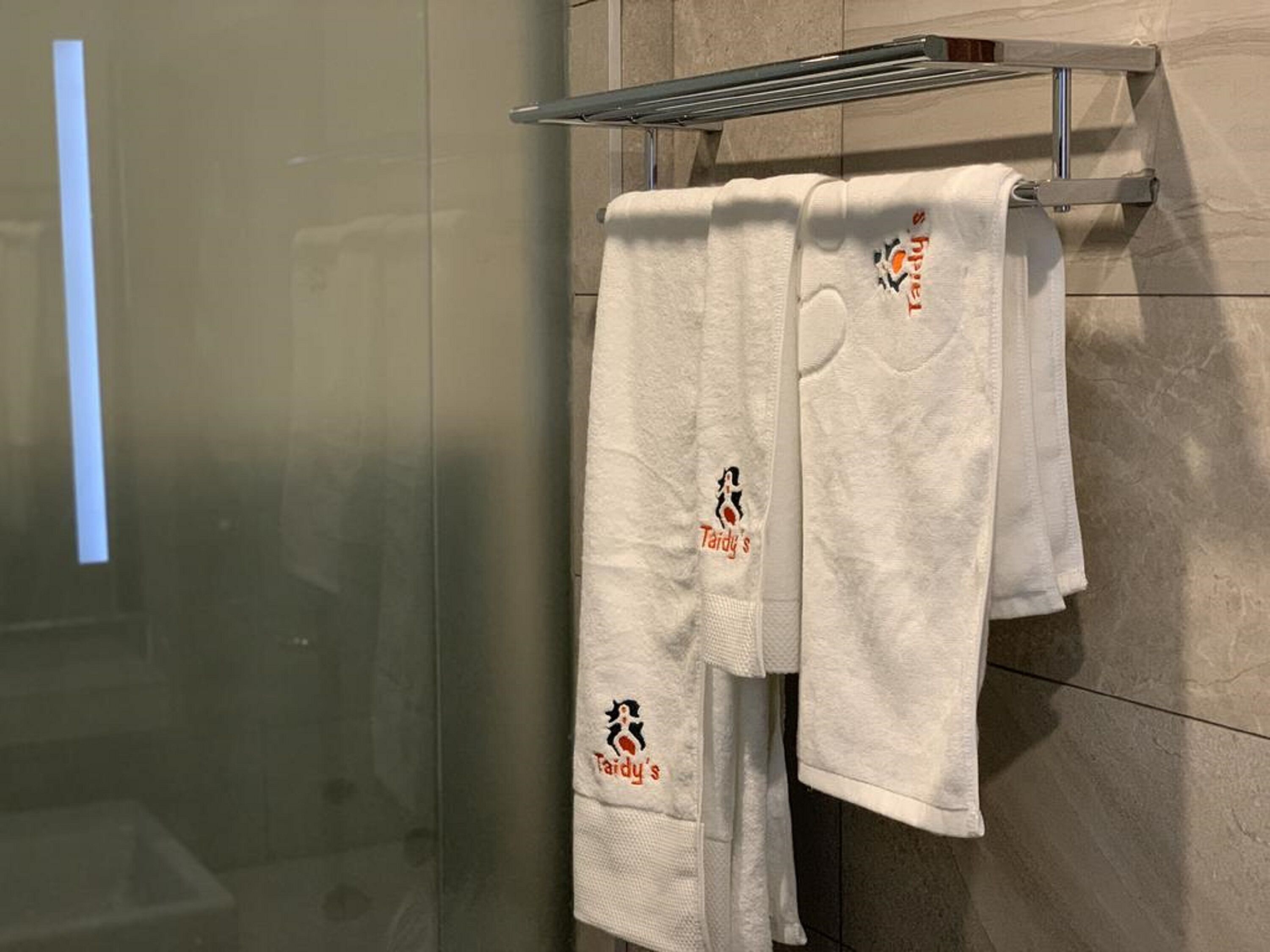 Un baño con mampara de vidrio, un perchero con dos toallas blancas y pared de azulejos.