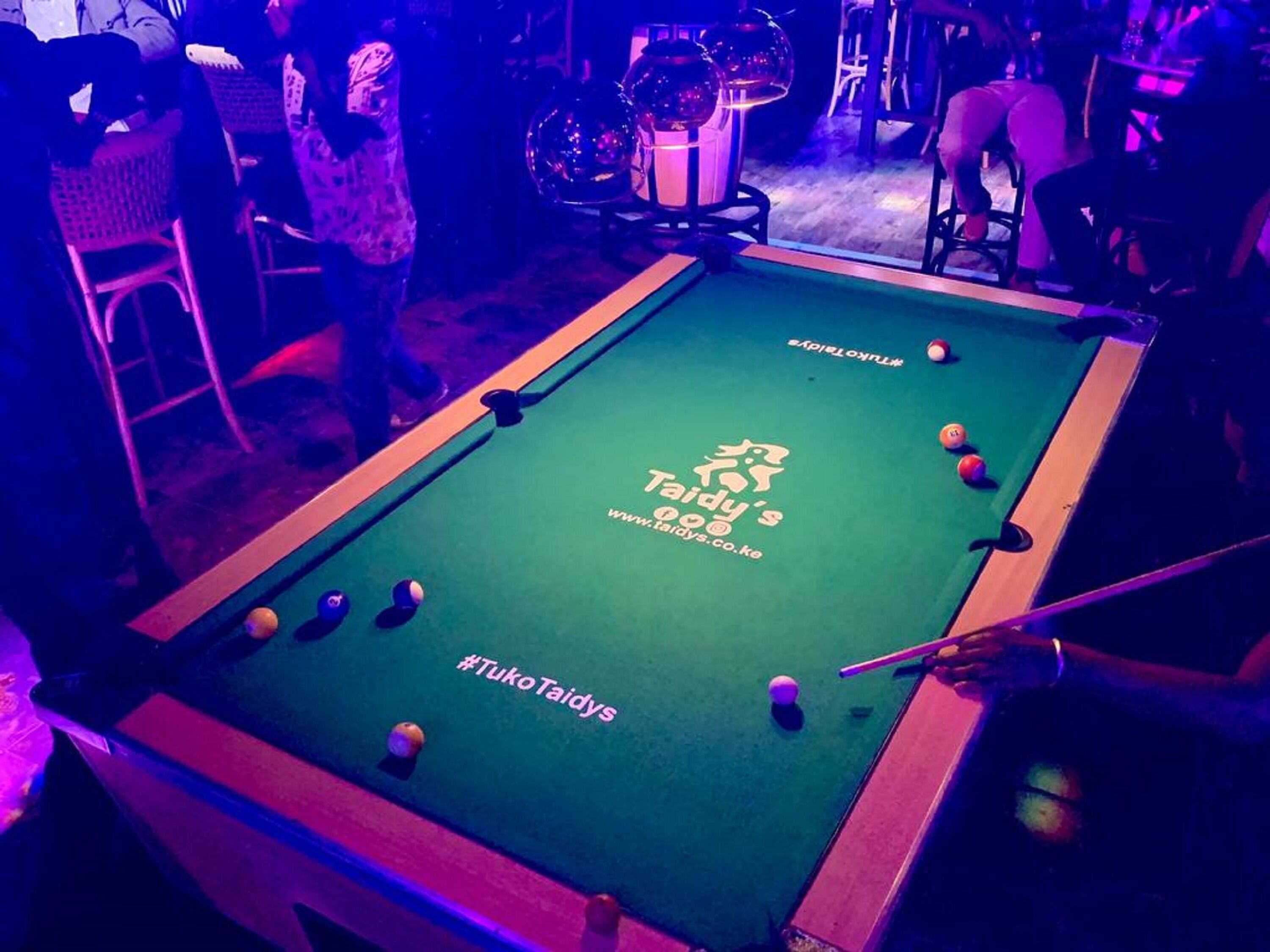 A pool table with a logo and text, surrounded by chairs and tables in a dimly lit room.
