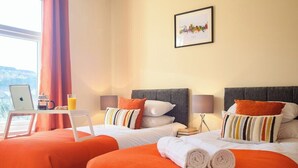 Hotellihuone, jossa on kaksi sänkyä, työpöytä kannettavalla tietokoneella, lasi appelsiinimehua ja kaupunkimaisemaa esittävä taulu seinällä.