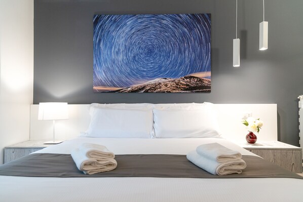 A hotel room with a large bed, white bedding, a nightstand with a lamp, a vase with flowers, and a star trail artwork on the wall.