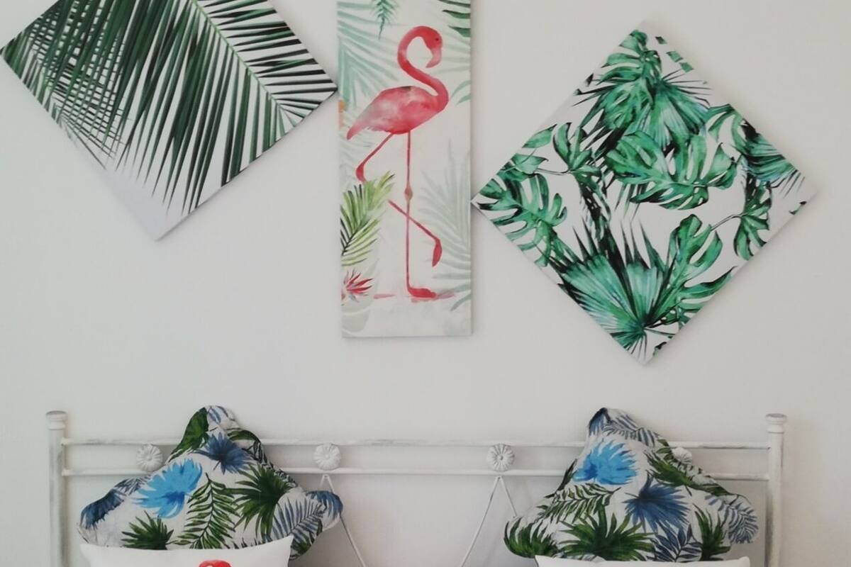 Ein Schlafzimmer mit einem Bett, zwei Kissen mit Flamingomotiven und drei Wandbildern mit tropischen Blättern und einem Flamingo.