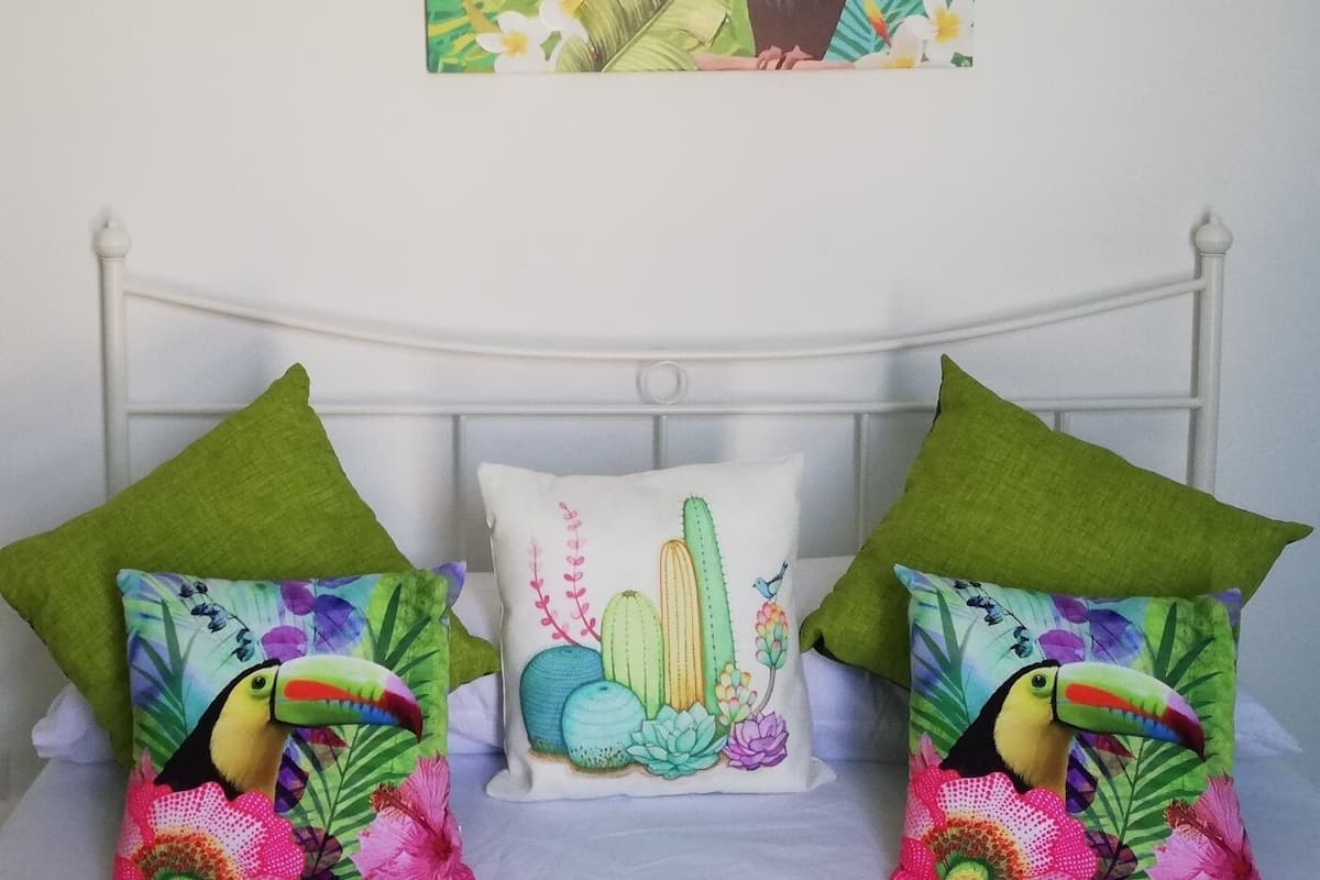 Ein Bett mit zwei grünen Kissen und einem weißen Kissen mit einem Kaktusmuster, sowie eine Wand mit einem Gemälde von Tukanen.
