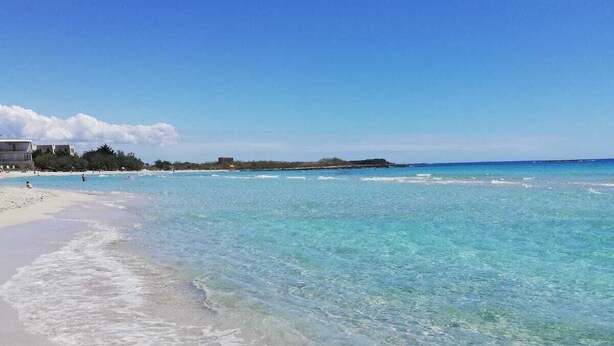 A beach with clear turquoise water, a sandy shore, and a building in the background. - Coast to coast Salento sea rooms (Porto Cesareo)