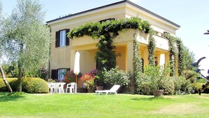 Un bâtiment à deux étages avec une façade jaune, un patio couvert doté de chaises blanches et d’une table, et un jardin avec différentes plantes et fleurs.