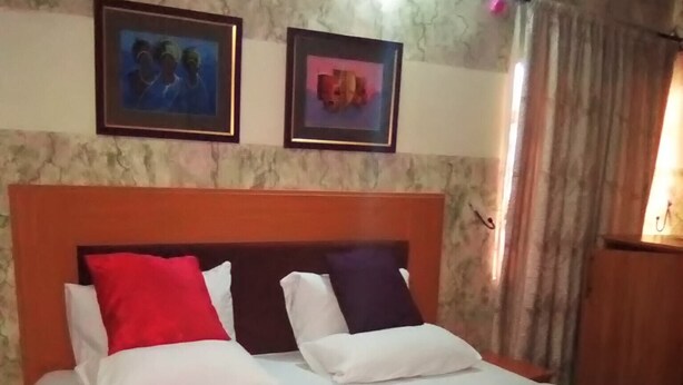 A neatly made bed with white bedding and two pillows, one red and one dark blue, in a room with a marble-patterned wall and two framed pictures above the bed.