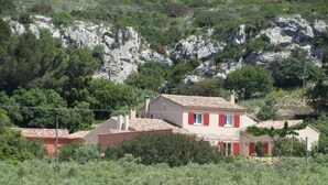 A two-story house with a red-tiled roof, surrounded by greenery and a rocky hillside.