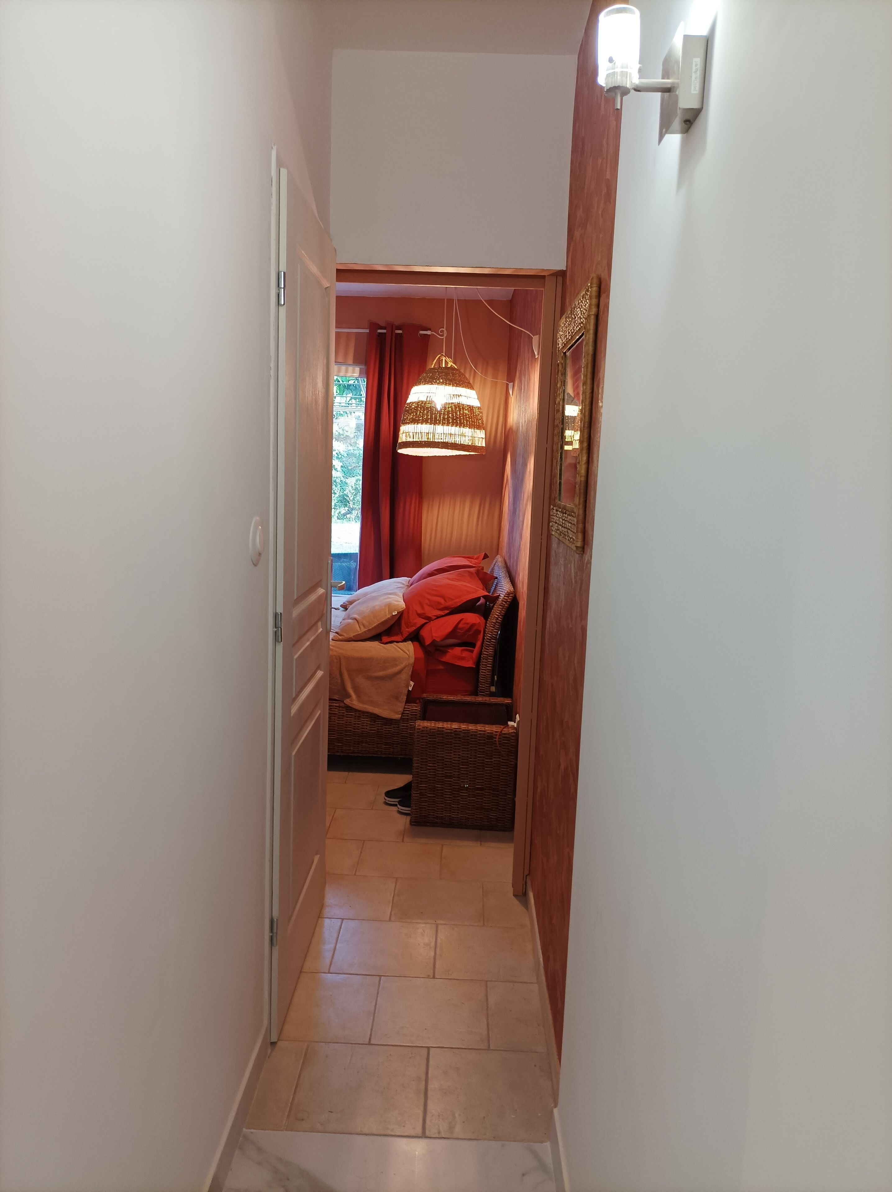 A narrow hallway leading to a bedroom with a bed, a hanging light fixture, and a wooden door.