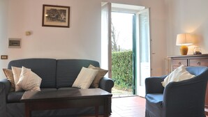 A living room with a sofa, armchair, and a table. There is a door leading outside, a framed picture on the wall, and a lamp on a side table.