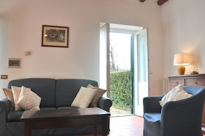 A living room with a sofa, armchair, and a table. There is a door leading outside, a framed picture on the wall, and a lamp on a side table. - Montecasciano Girasole (Capranica)