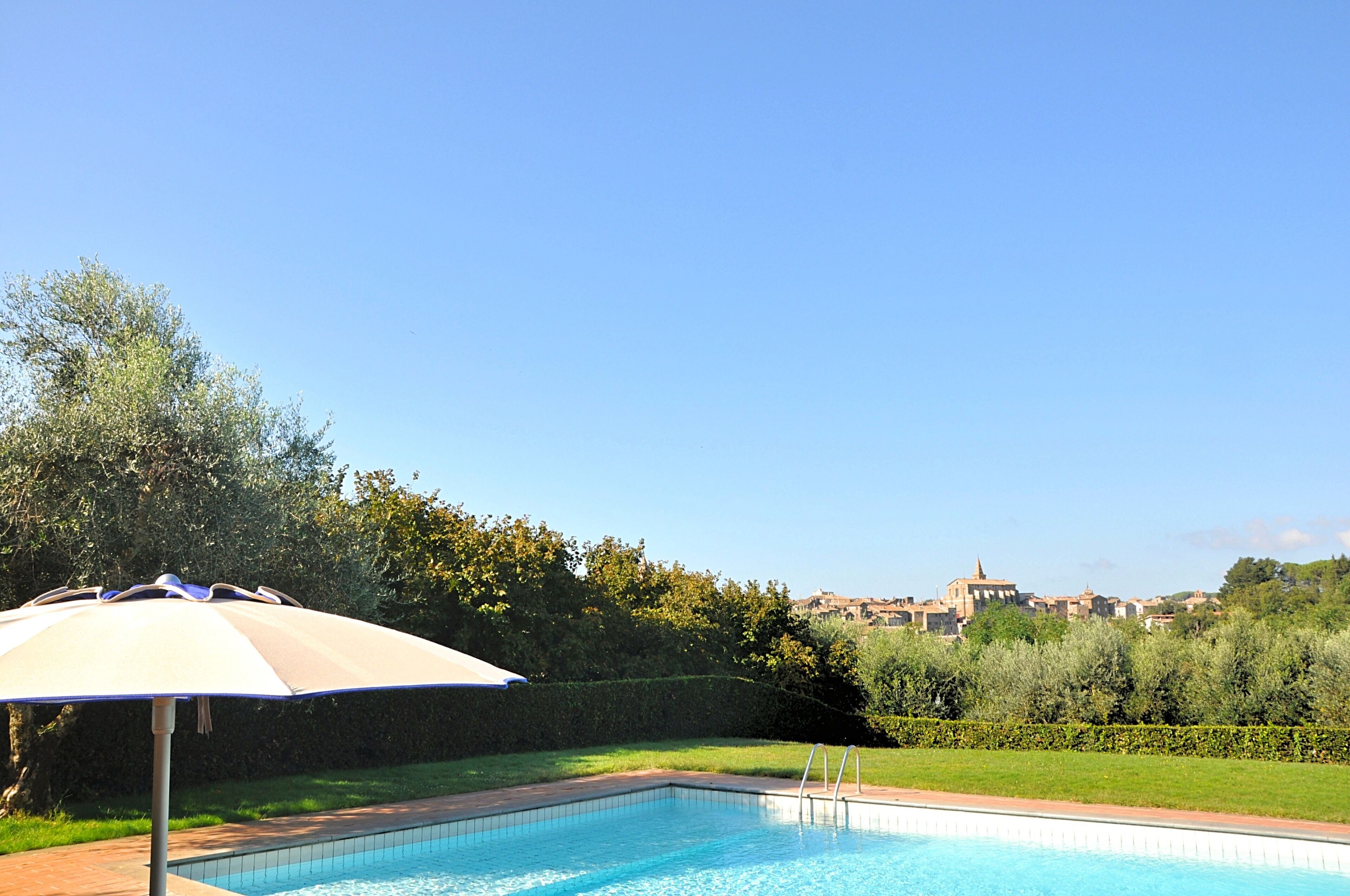A swimming pool with a sun umbrella, surrounded by greenery and a view of distant buildings.