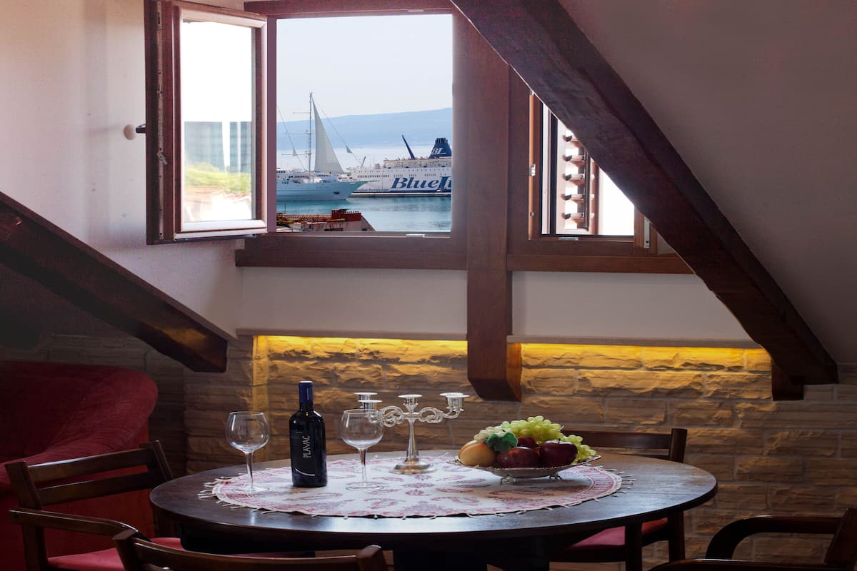 Un coin repas avec une table dressée pour deux personnes, une bouteille de vin, des verres à vin et un panier de fruits. Une fenêtre offre une vue sur des bateaux et un voilier sur l’eau.