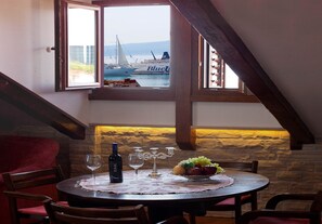 A dining area with a table set for two, a bottle of wine, wine glasses, and a fruit bowl. A window offers a view of boats and a sailboat on the water.