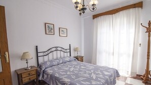 Une chambre avec un sol à carreaux, un lit recouvert d’une couvre-lit à motifs floraux, deux tables de chevet avec des lampes, un porte-manteau en bois et un lustre.