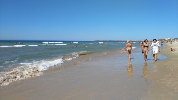 Une plage où l’on voit des gens se promener le long du rivage, des vagues qui se brisent sur le sable et un ciel bleu dégagé.