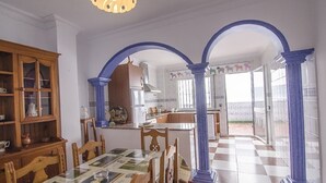 Un espace repas avec un sol à carreaux, des chaises en bois et une table recouverte d’une nappe imprimée. Des colonnes bleues encadrent l’espace, et on aperçoit un coin cuisine au fond.
