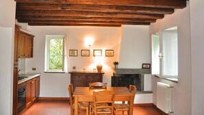 A kitchen with wooden cabinets, a dining table, chairs, a fireplace, and a window with a view of greenery. - Montecasciano Oleandro (Capranica)