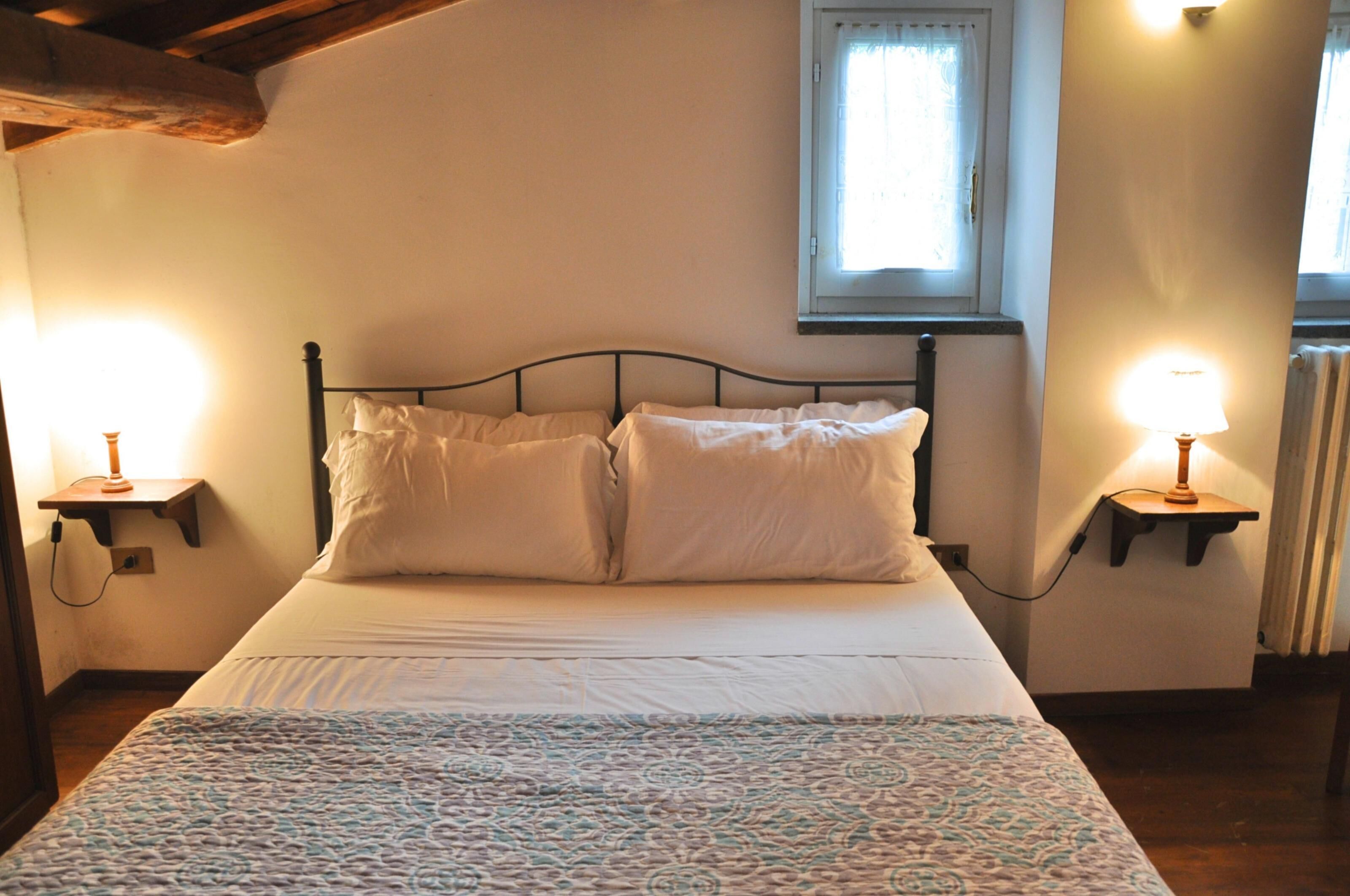 A neatly made bed with white linens, two bedside lamps, and a window with curtains.