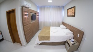A hotel room with a bed, a bedside table with a phone and remote, a wardrobe, a TV, and a painting on the wall.