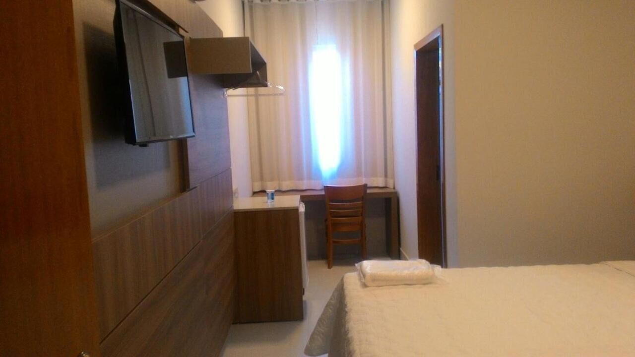 Habitación de hotel con cama, escritorio, silla, televisor montado en la pared y ventana con cortinas.