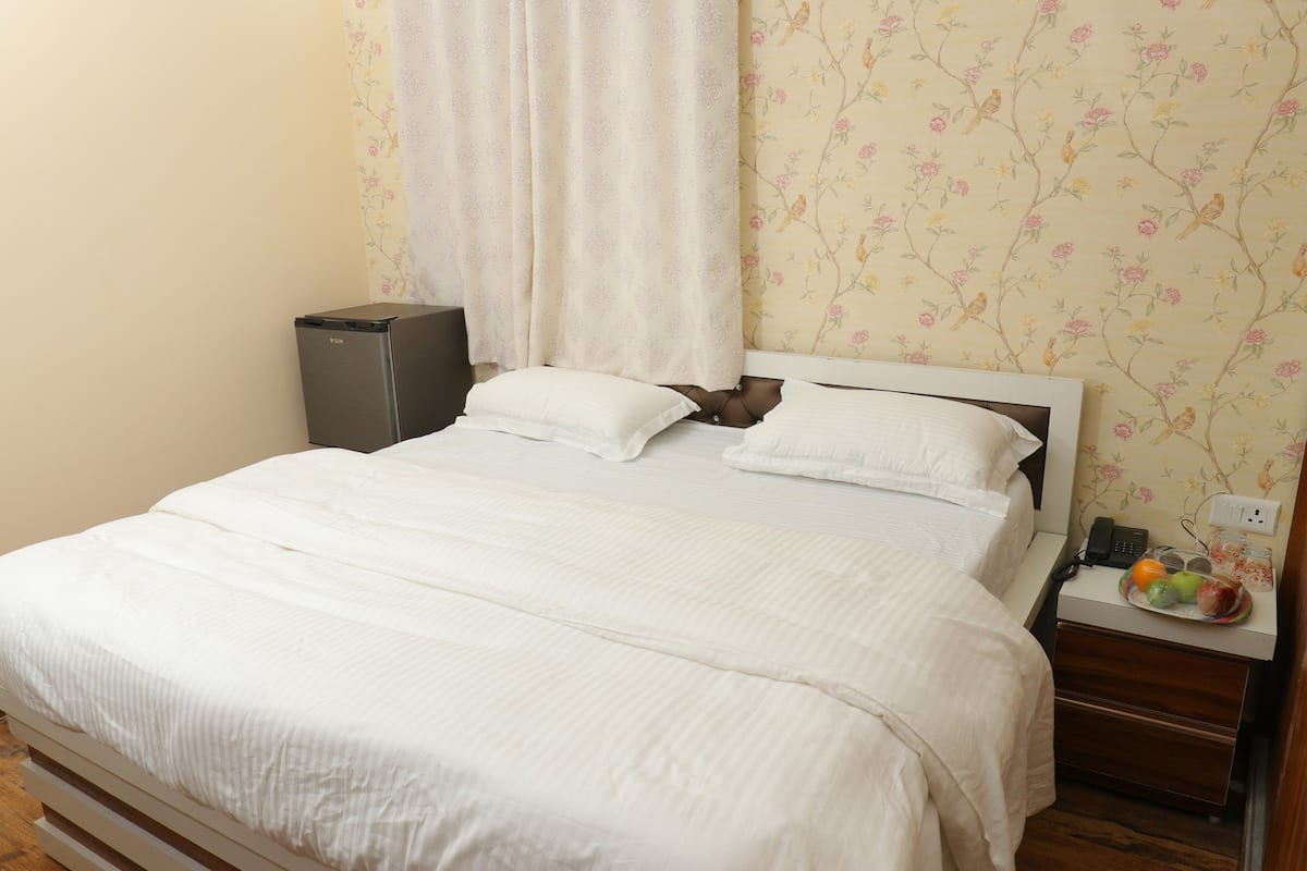 A neatly made bed with white bedding, a bedside table with a phone and fruit, and a small speaker on the nightstand.
