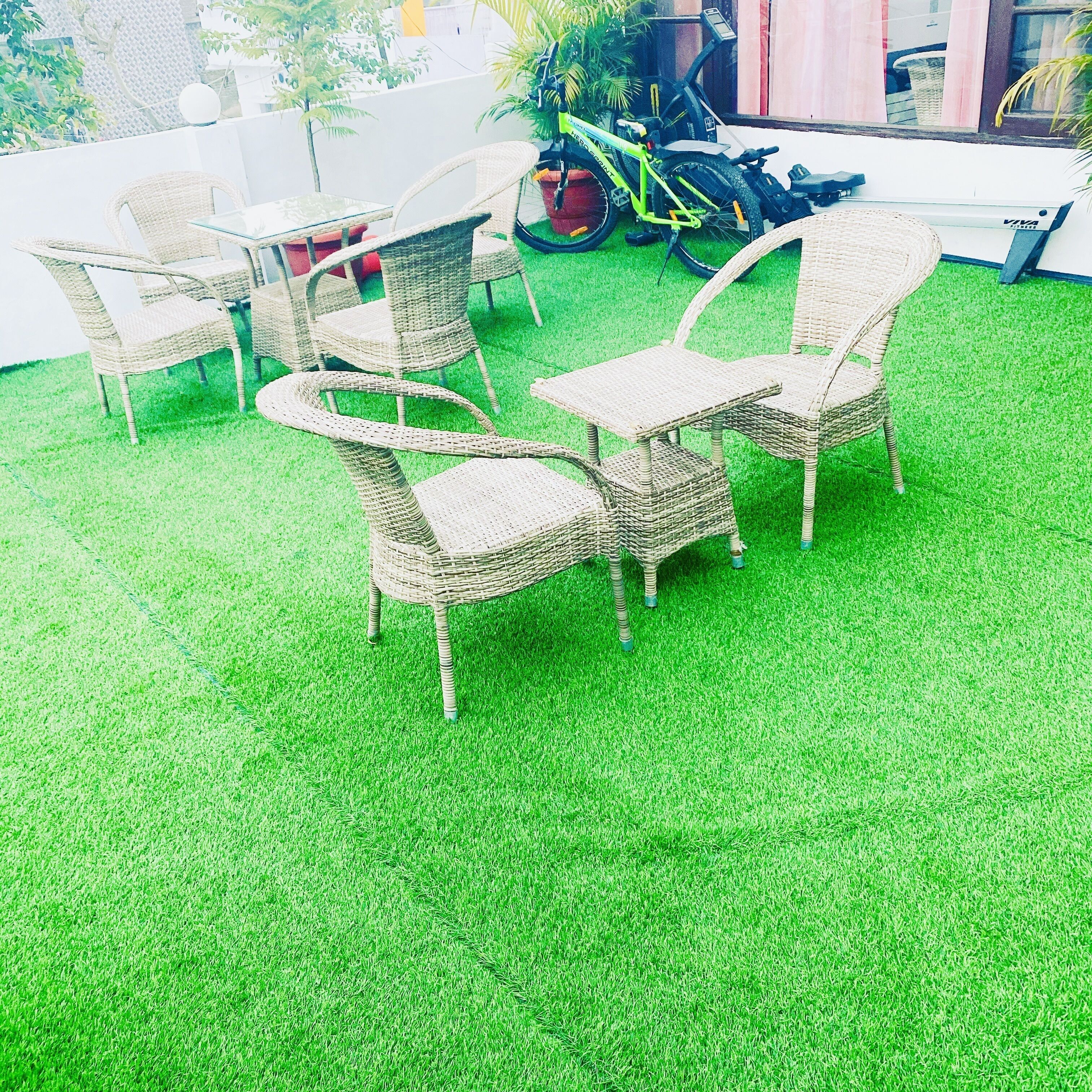 Wicker chairs and a table on artificial grass, with a bicycle and a scooter in the background.
