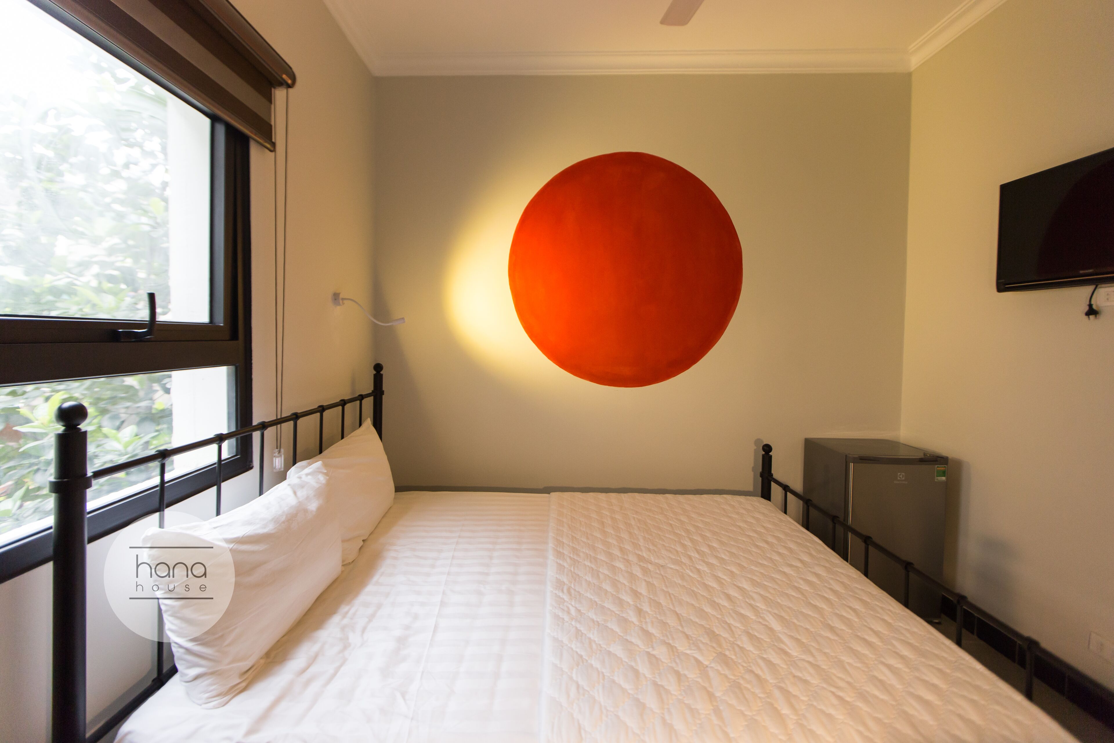 A bedroom with a bed, a window, a wall-mounted TV, and a refrigerator.