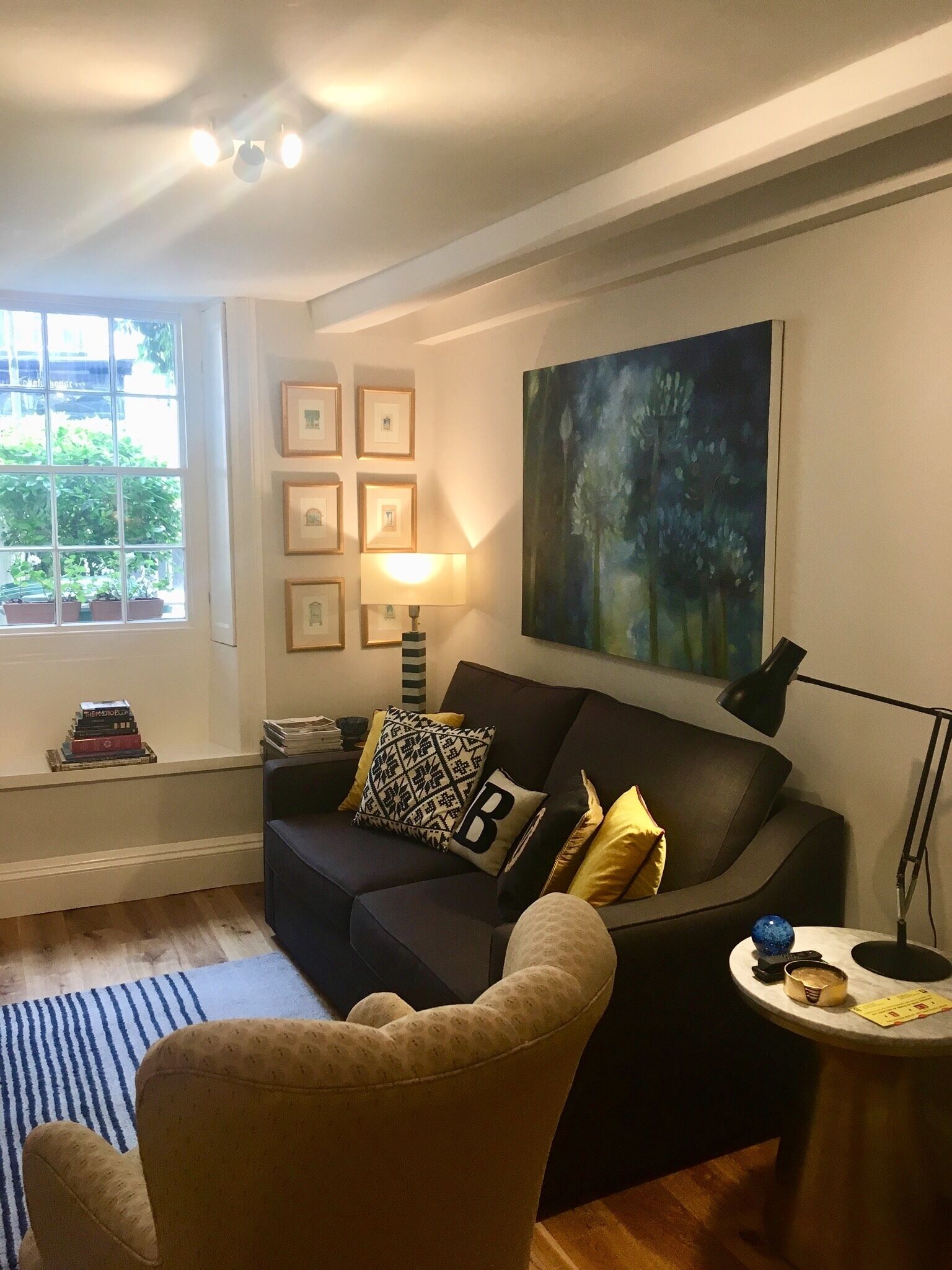 A living room with a dark-colored sofa, a patterned armchair, a side table with a lamp, and a large abstract painting on the wall.