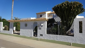 A white building with a large cactus in front, a palm tree, and a clear blue sky.
