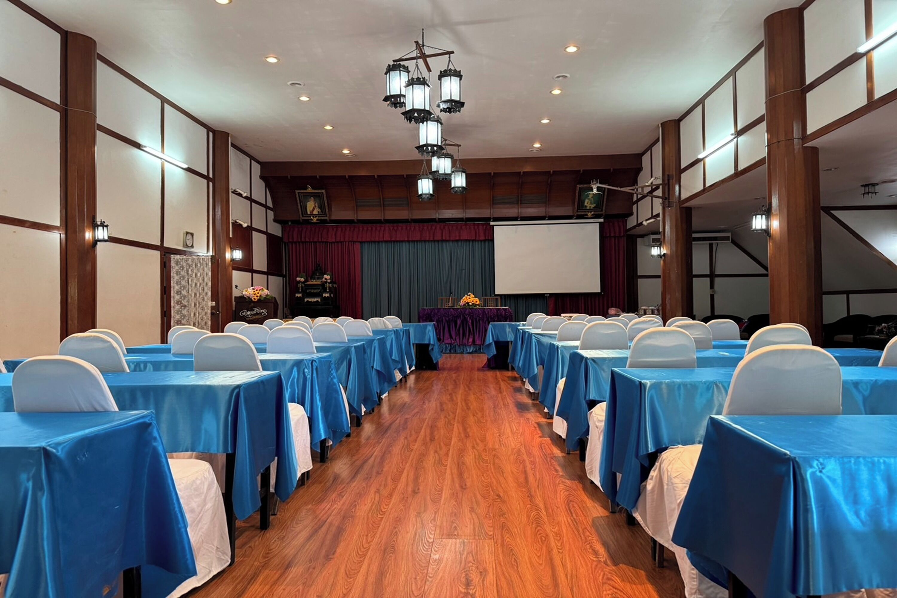 Un salón de conferencias con filas de mesas y sillas, paneles de madera y un escenario con una pantalla.