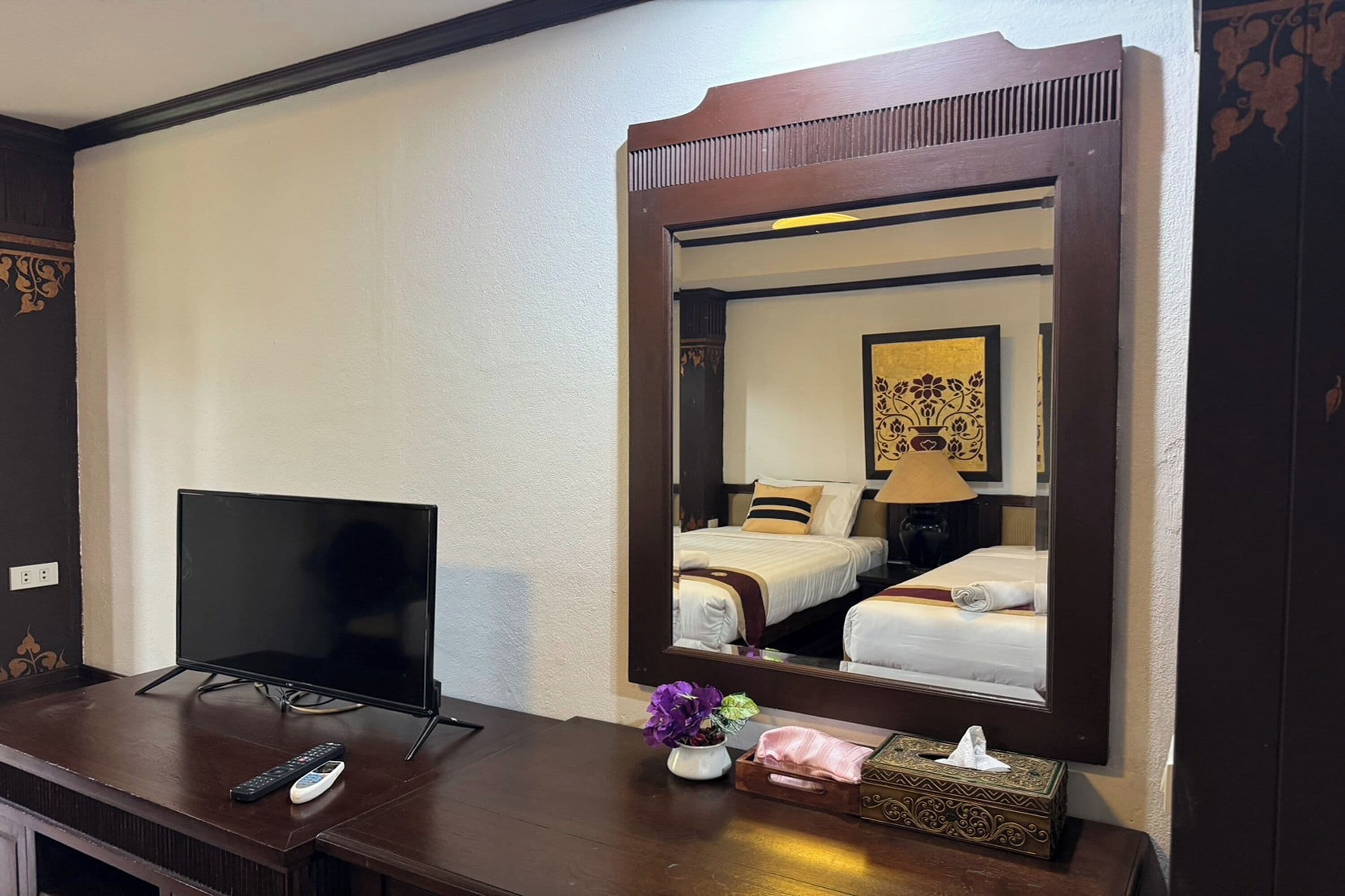 Habitació d'hotel amb un televisor sobre una taula de fusta, un mirall que reflecteix un llit i una làmpada, i un ram de flors en un vas petit.