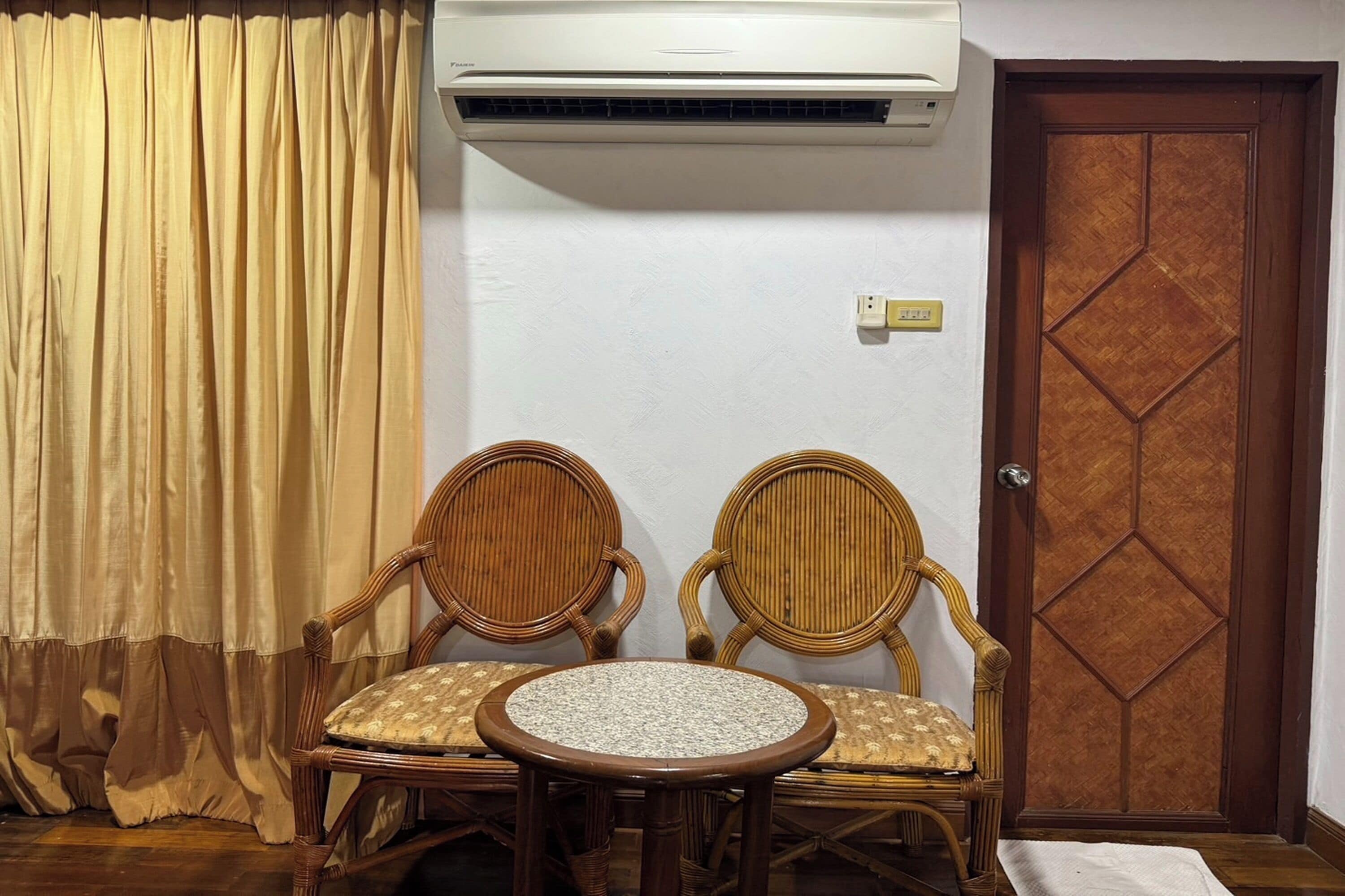 A room with two wicker chairs, a small round table, an air conditioning unit, a wooden door, and a curtain.