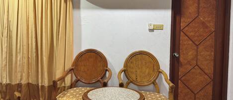 A room with two wicker chairs, a small round table, an air conditioning unit, a wooden door, and a curtain.