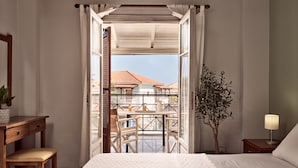 Une chambre avec un grand lit, un bureau en bois, une chaise, une table de chevet, une lampe et une vue sur un balcon aménagé avec du mobilier d’ext érieur.